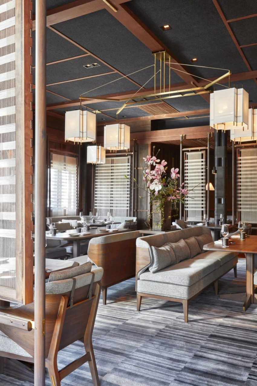 Modern restaurant interior with wooden furnishings, elegant lighting, and a central floral arrangement on a striped carpet.
