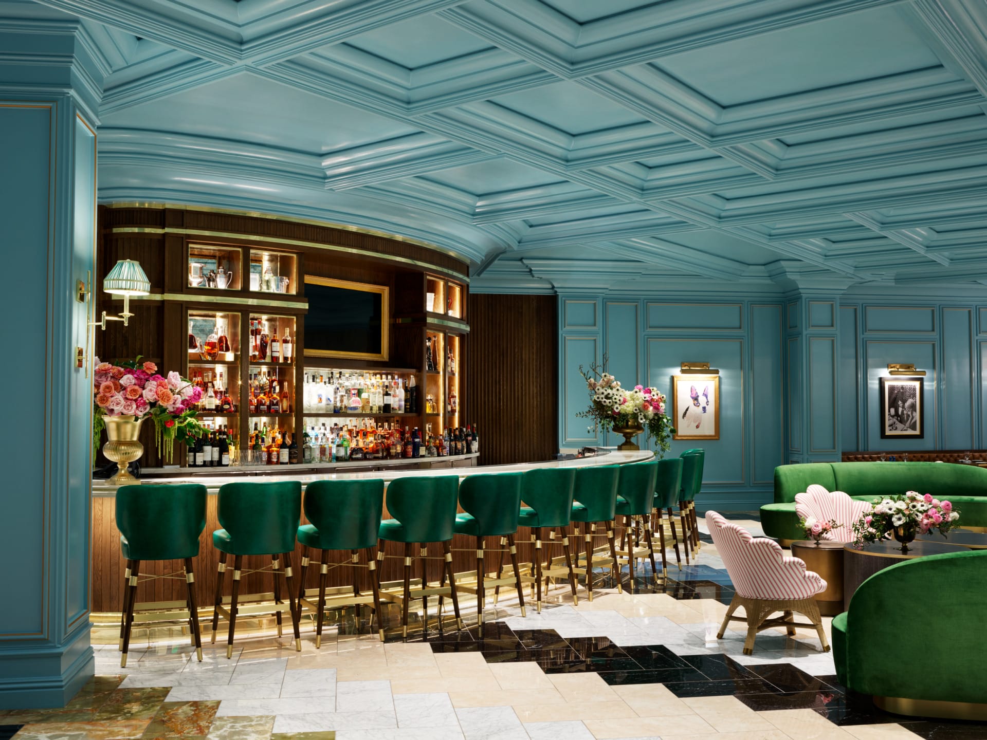 Elegant bar interior with teal walls, green velvet barstools, and a marble counter adorned with floral arrangements.