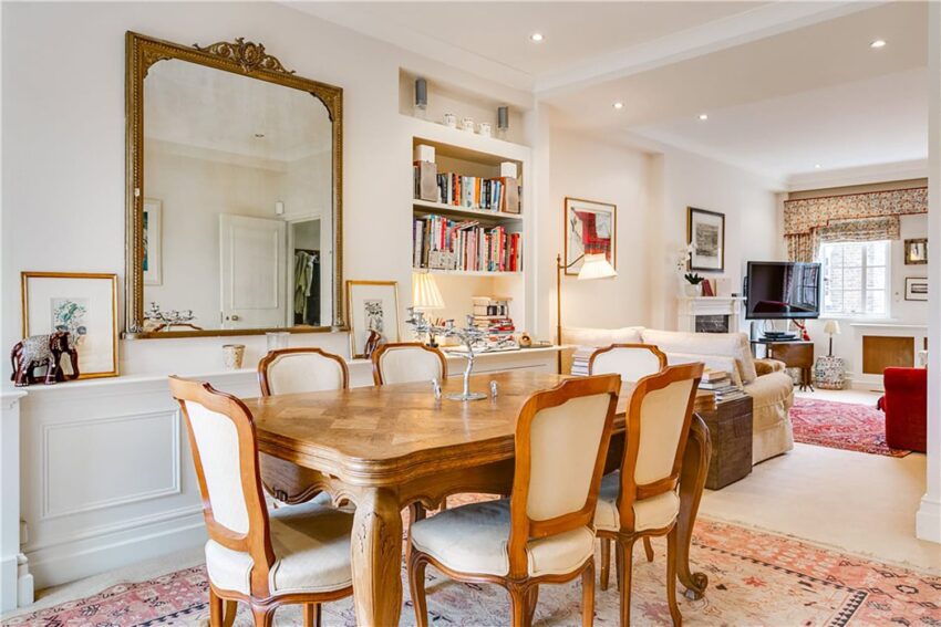 Elegant dining room with a wooden table, vintage chairs, large mirror, bookshelves, and cozy living area in the background.