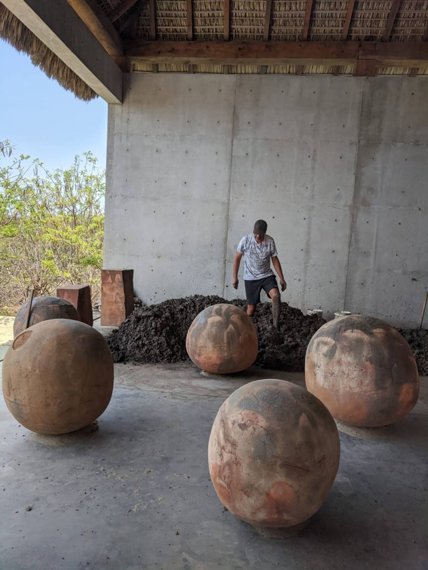 Person working with large clay spheres under a thatched roof, surrounded by greenery and concrete walls.
