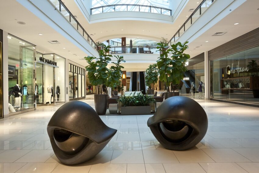 Modern shopping mall interior with stylish black sculptures and lush green plants, showcasing various fashion stores.