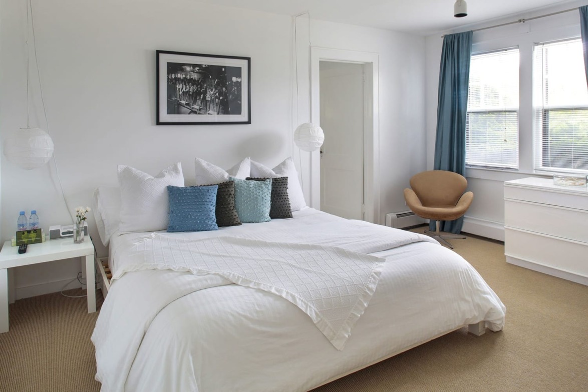 Modern bedroom with white bedding, colorful pillows, a bedside table with water bottles, and a window with blue curtains.