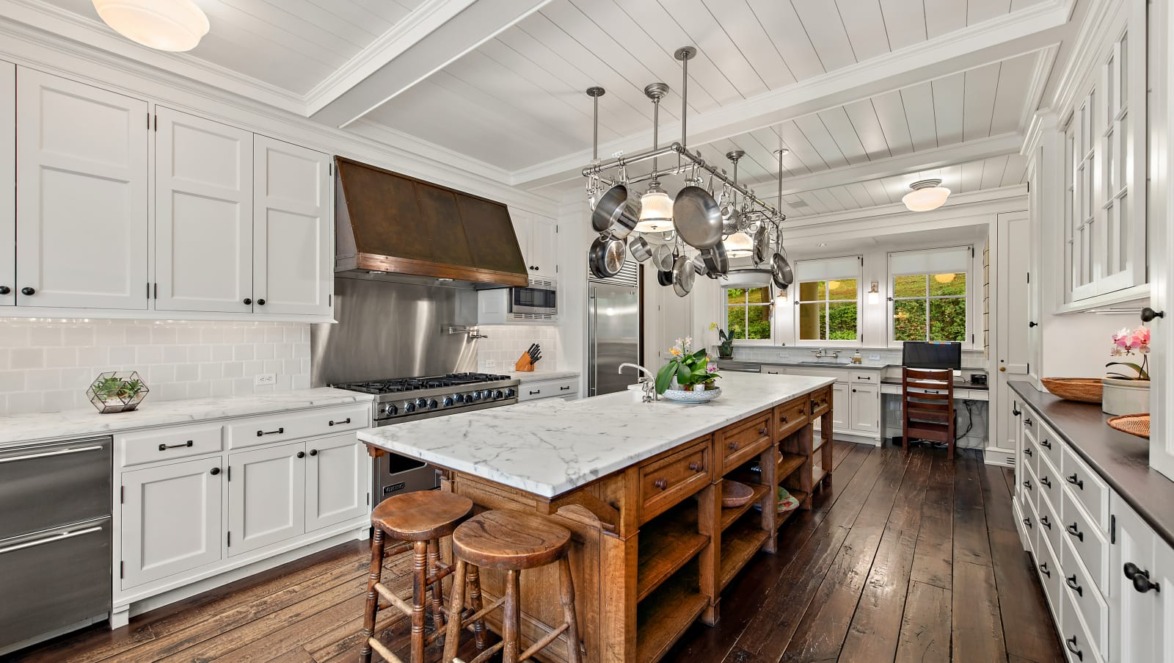 Spacious kitchen with white cabinets, wooden island, marble countertop, hanging pots, and stools on wooden floor.