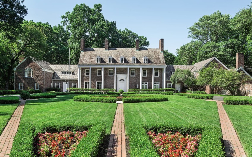 Large brick mansion with tall columns, surrounded by lush greenery and manicured gardens under a clear blue sky.