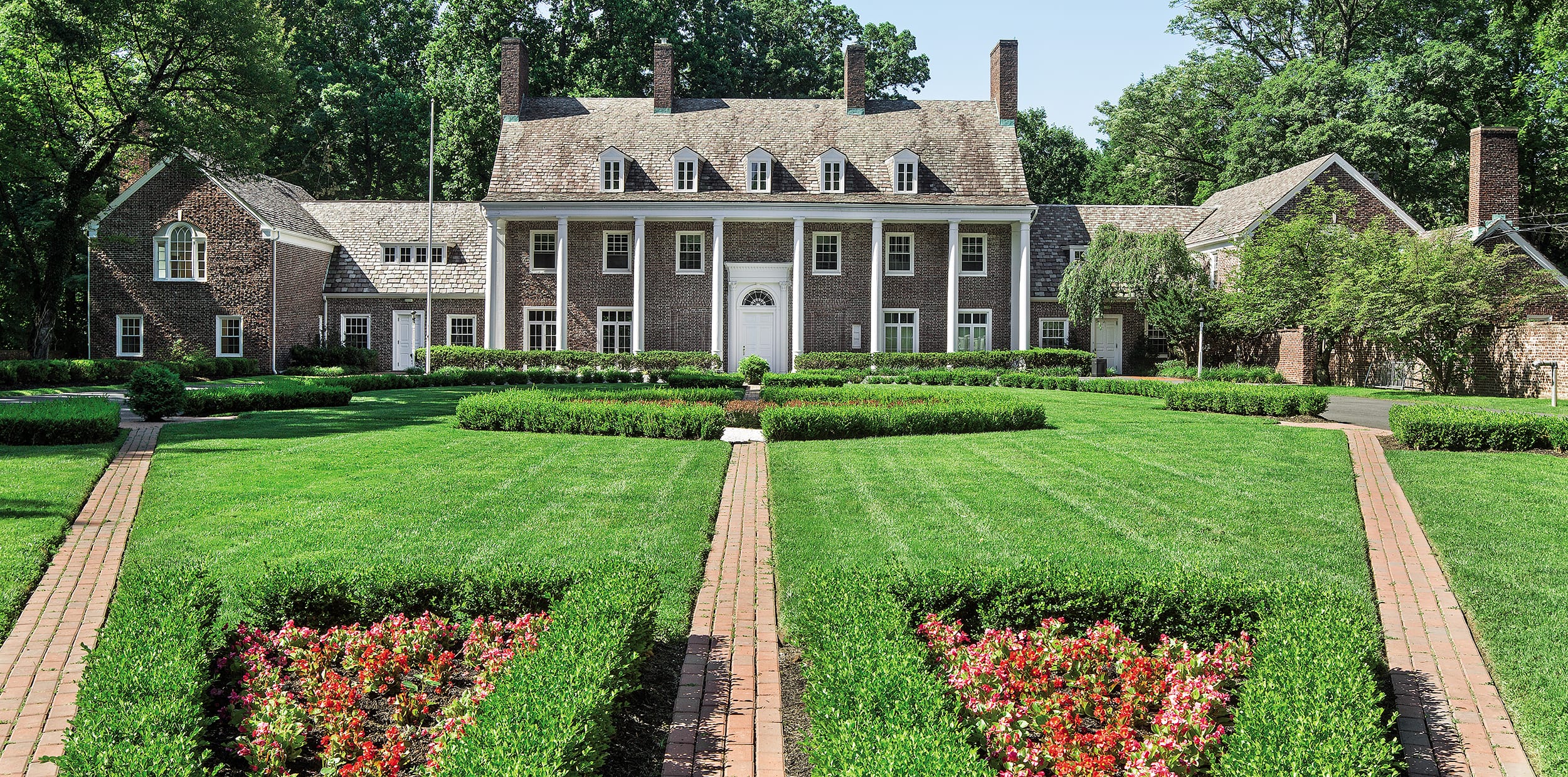 Large brick mansion with white columns, surrounded by manicured lawns, colorful flowers, and a symmetrical garden pathway.