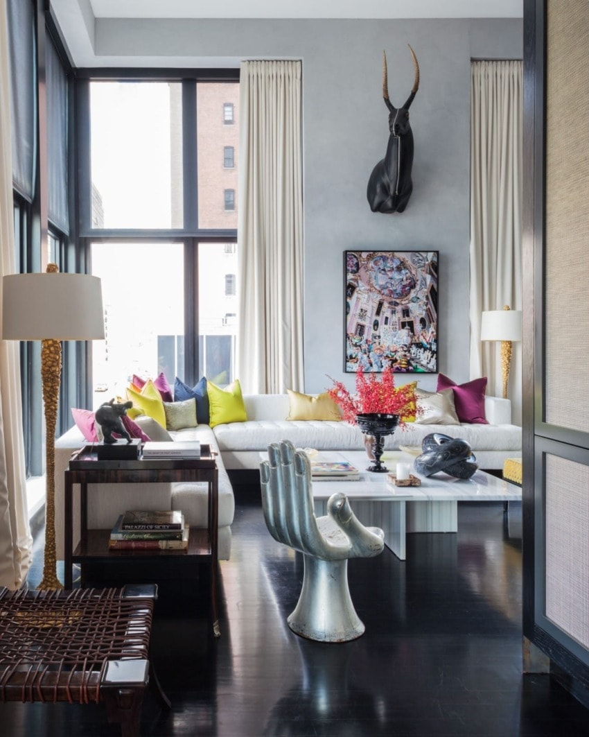 Modern living room with large windows, colorful pillows, unique hand chair, and elegant decor featuring a wall-mounted sculpture.