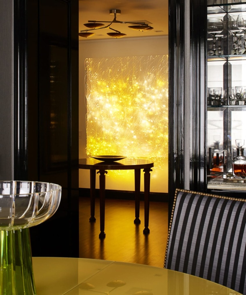 Dining area with a modern light installation glowing yellow, surrounded by dark furniture and a table with glassware.