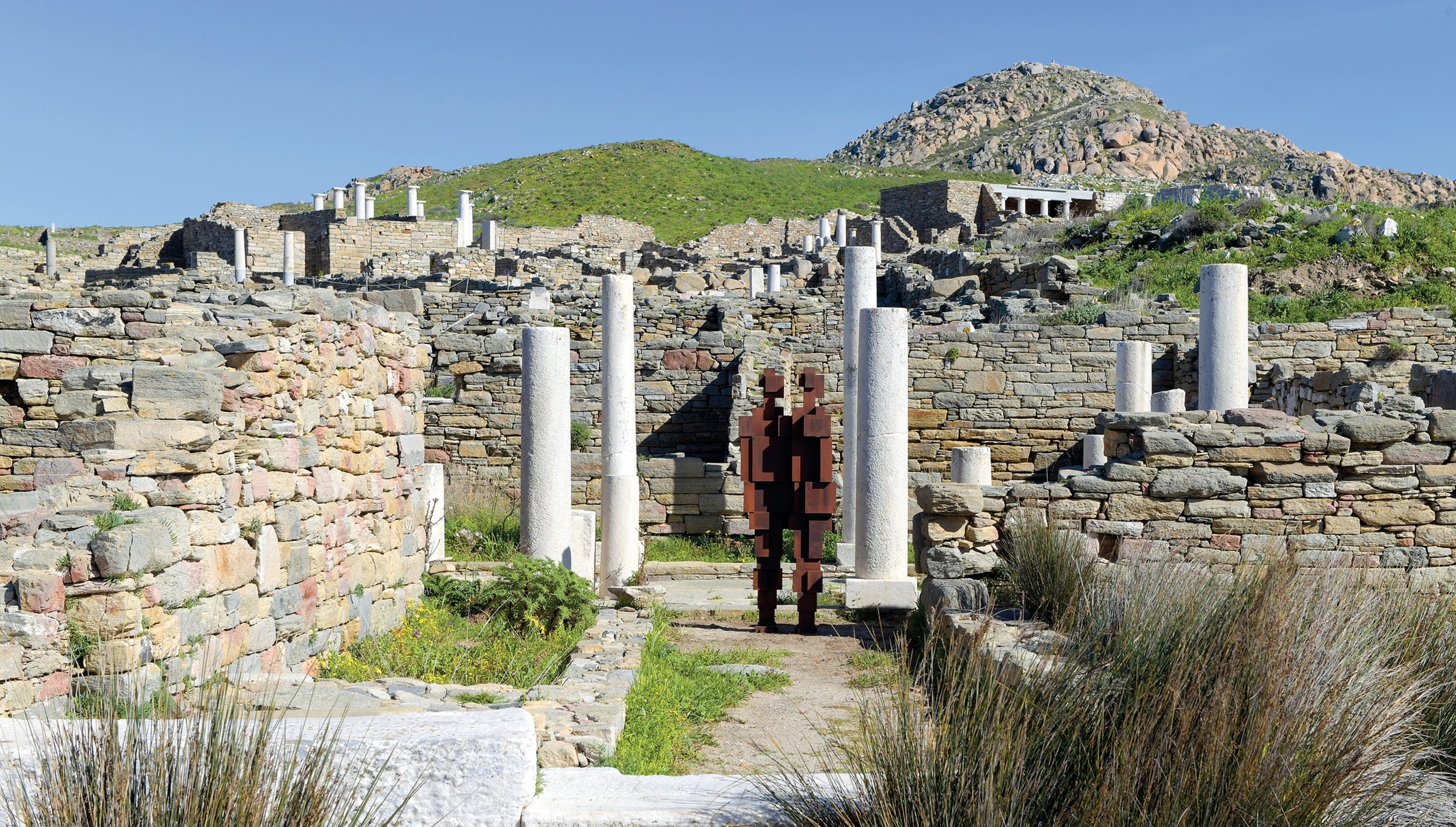 Ancient ruins with stone columns and walls under a clear blue sky, grassy hill in the background.