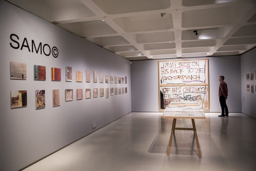 Art gallery with various paintings on walls, person observing large artwork, SAMO© text visible on left wall.