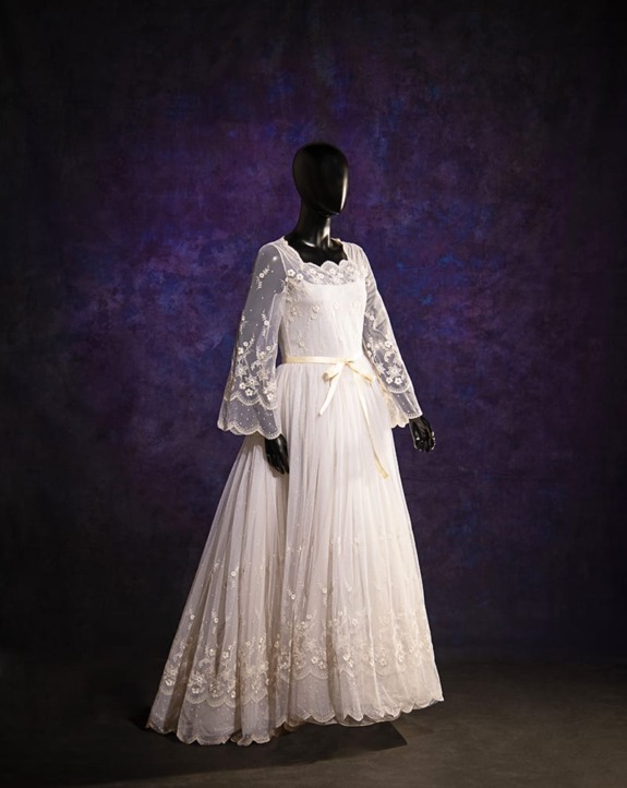 Mannequin displaying a vintage white lace wedding dress with bell sleeves and a bow, set against a dark textured background.