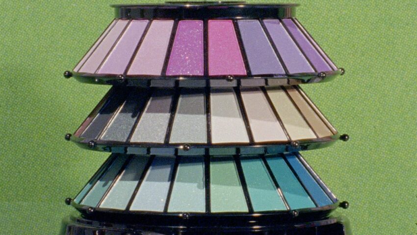 Tiered display of colorful eyeshadow palettes with purple, pink, gray, and teal shades on a green textured background.