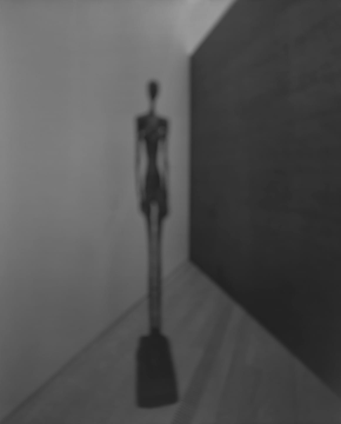 Blurry abstract sculpture of a tall, slender figure in a minimalist gallery with dark and light walls.