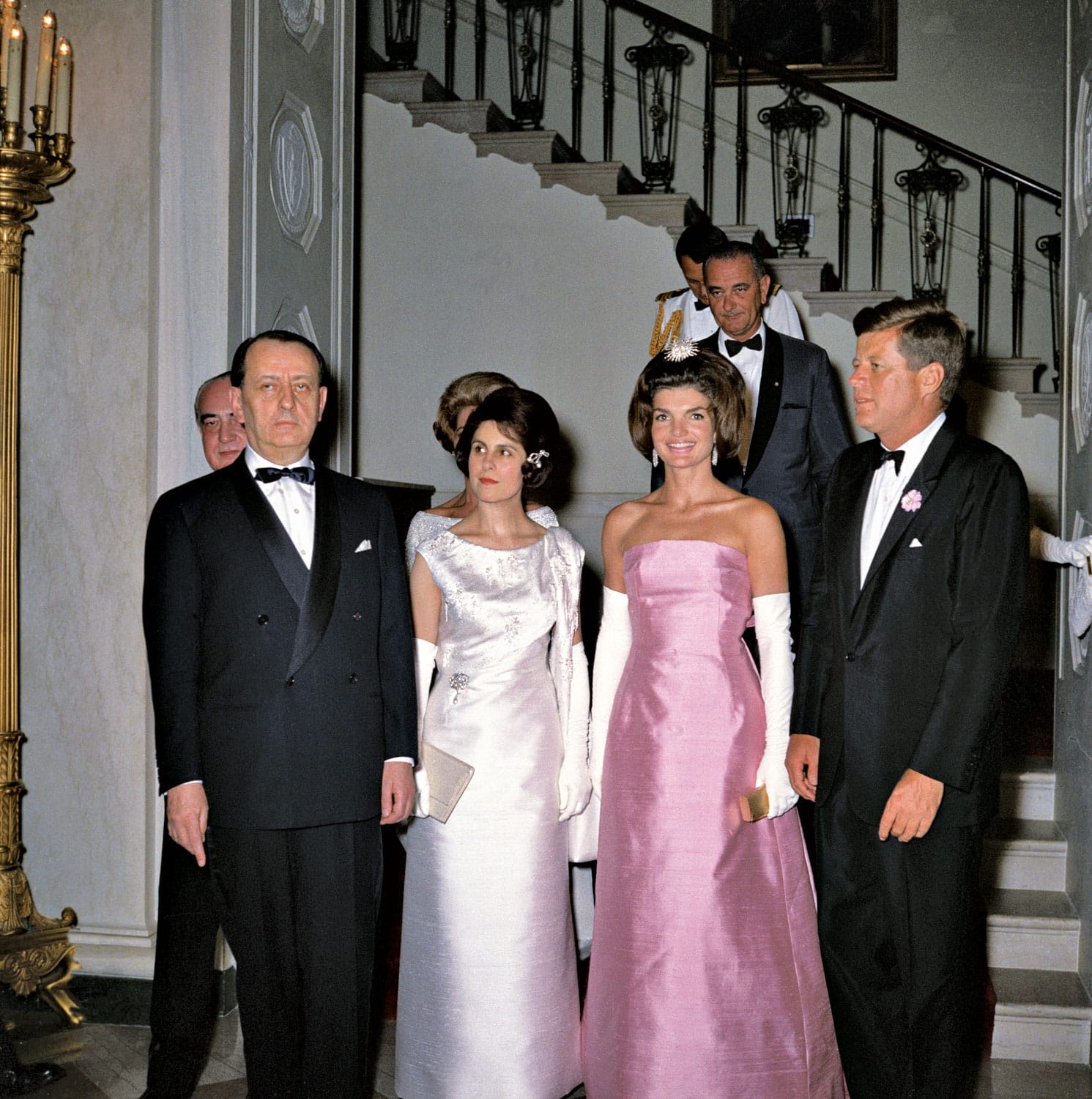 Group of elegantly dressed people standing in an ornate room with a staircase in the background.