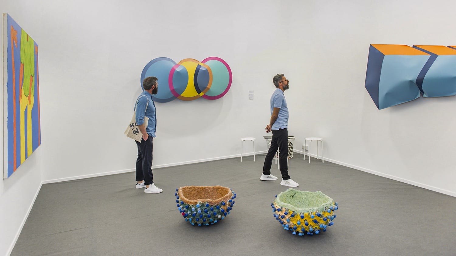 Two people observe modern art installations in a gallery, featuring colorful abstract wall pieces and textured floor sculptures.