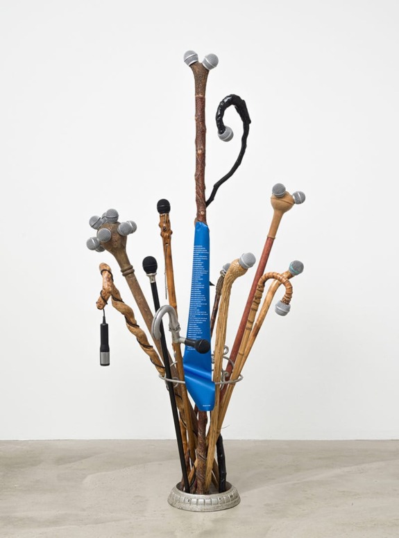 Collection of walking sticks in a stand, some with unique handles resembling microphones, against a plain white background.