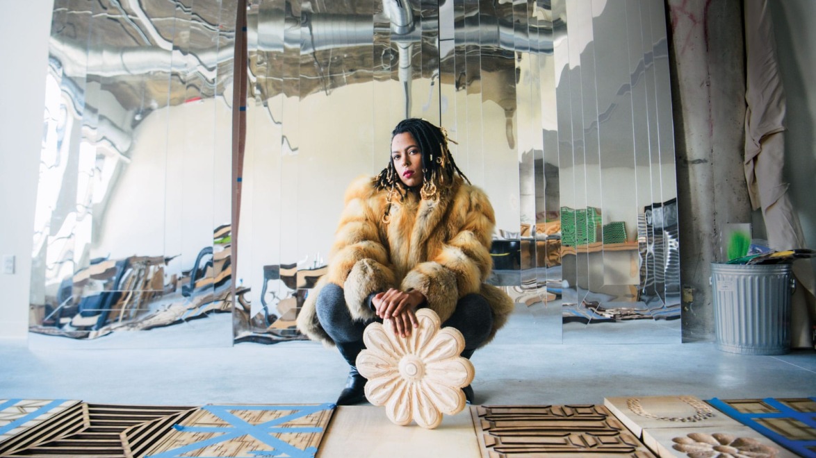 Person in fur coat squatting in art studio with reflective wall and wooden carvings on the floor