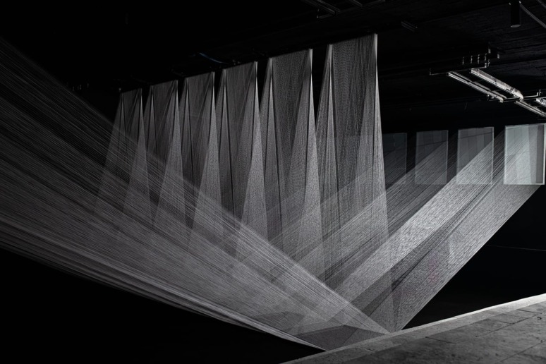 Installation art with diagonal strings creating a geometric pattern in a dark room, illuminated by overhead lights.