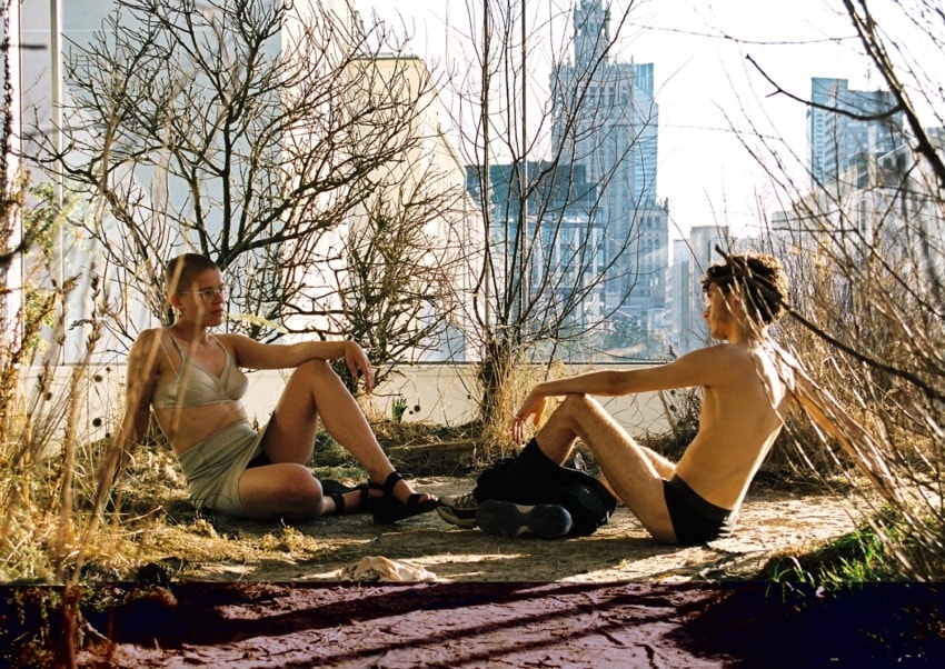 Two people relaxing outdoors among dry plants with urban buildings in the background.