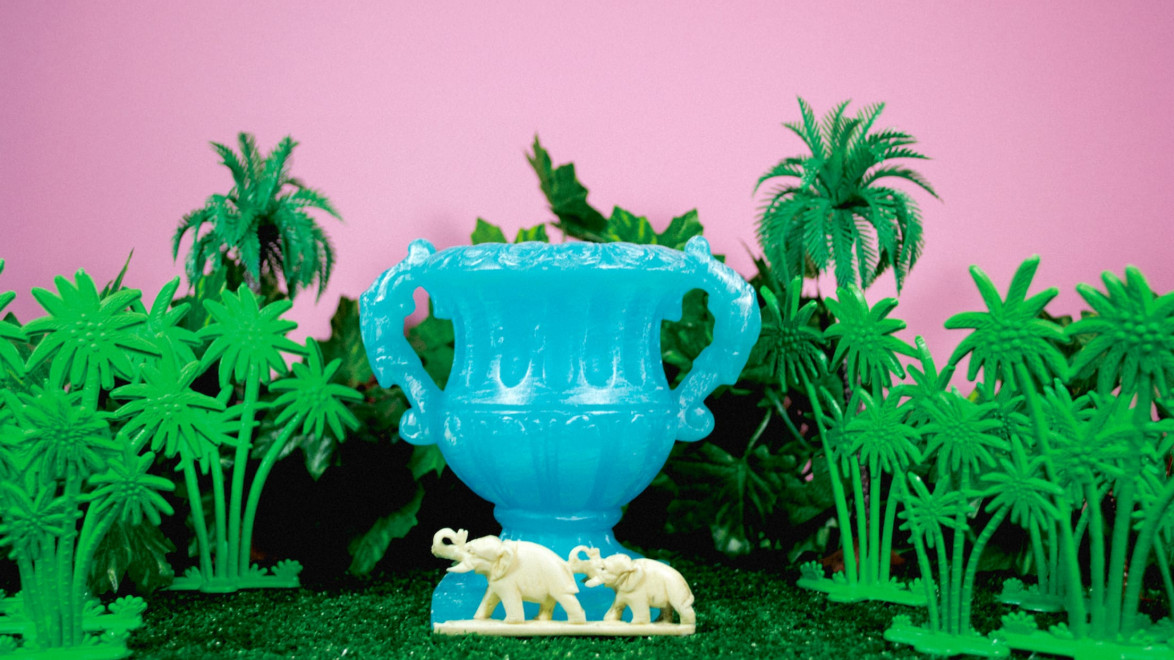 Blue vase surrounded by toy palm trees and two white toy elephants on a grassy surface against a pink background
