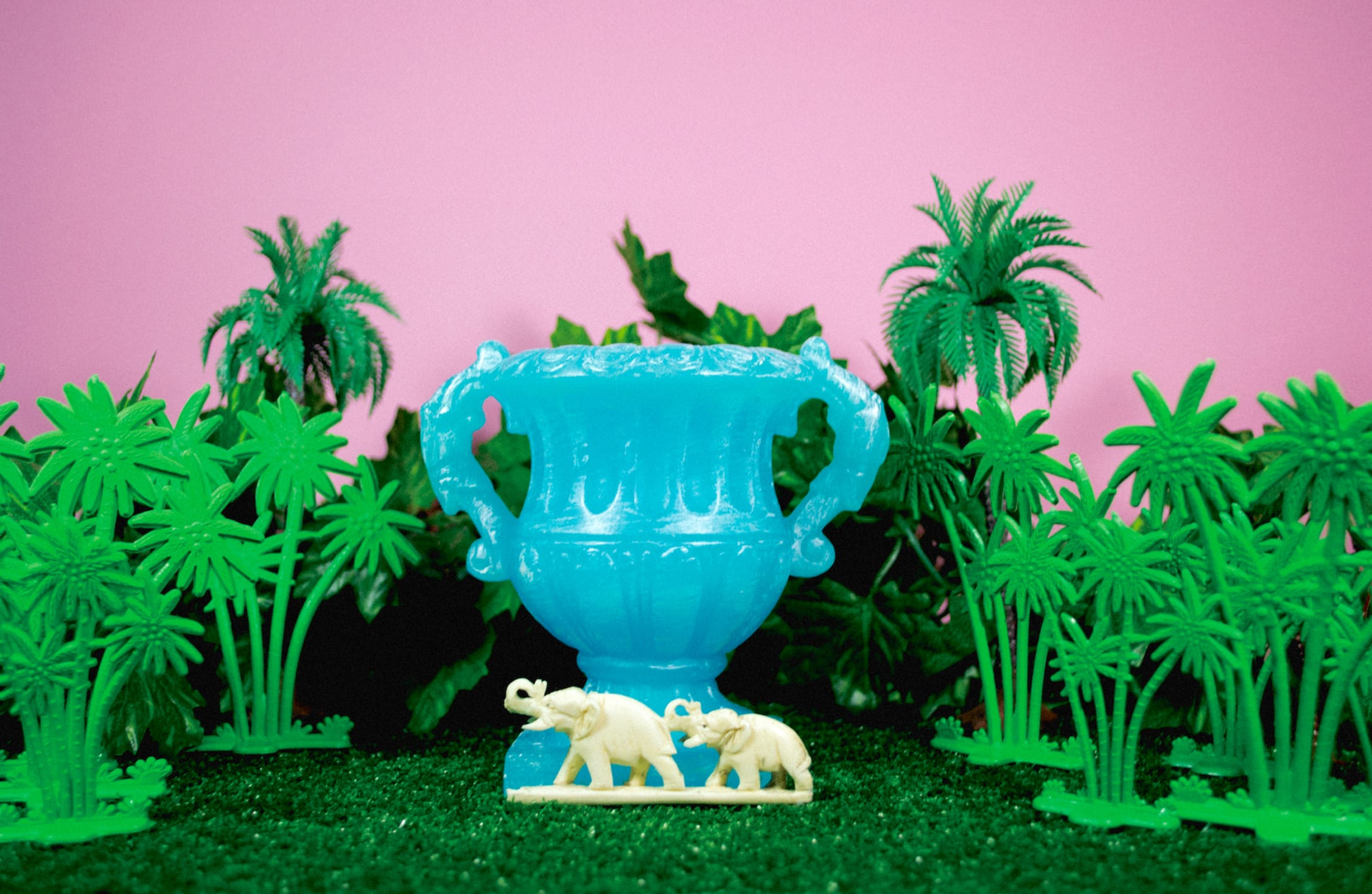 Blue vase surrounded by toy palm trees and two white toy elephants on a grassy surface against a pink background