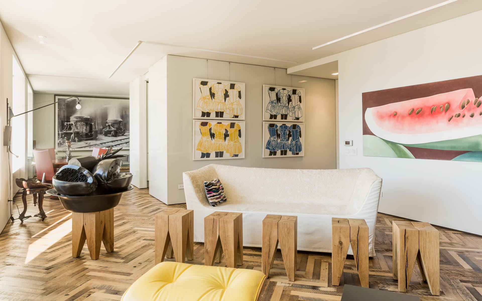 Modern living room with abstract artwork, white sofa, wooden stools, and parquet flooring. Bright and spacious interior design.