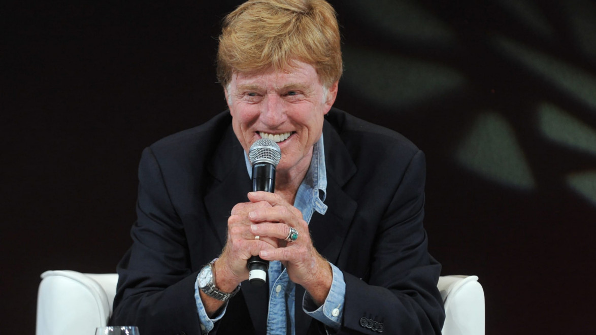 Man smiling while holding a microphone, wearing a black jacket and light blue shirt, seated on a white chair on stage.