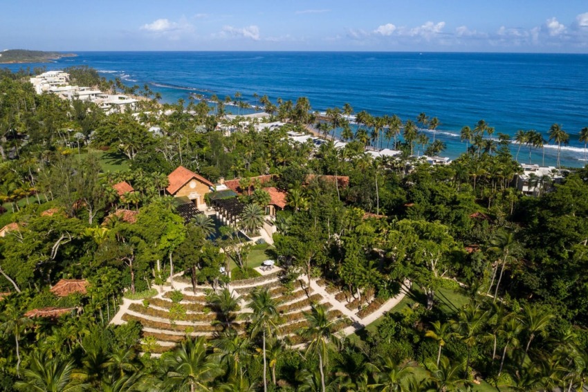 Aerial view of a coastal resort with lush greenery, palm trees, and a shoreline meeting the clear blue ocean under a sunny sky.