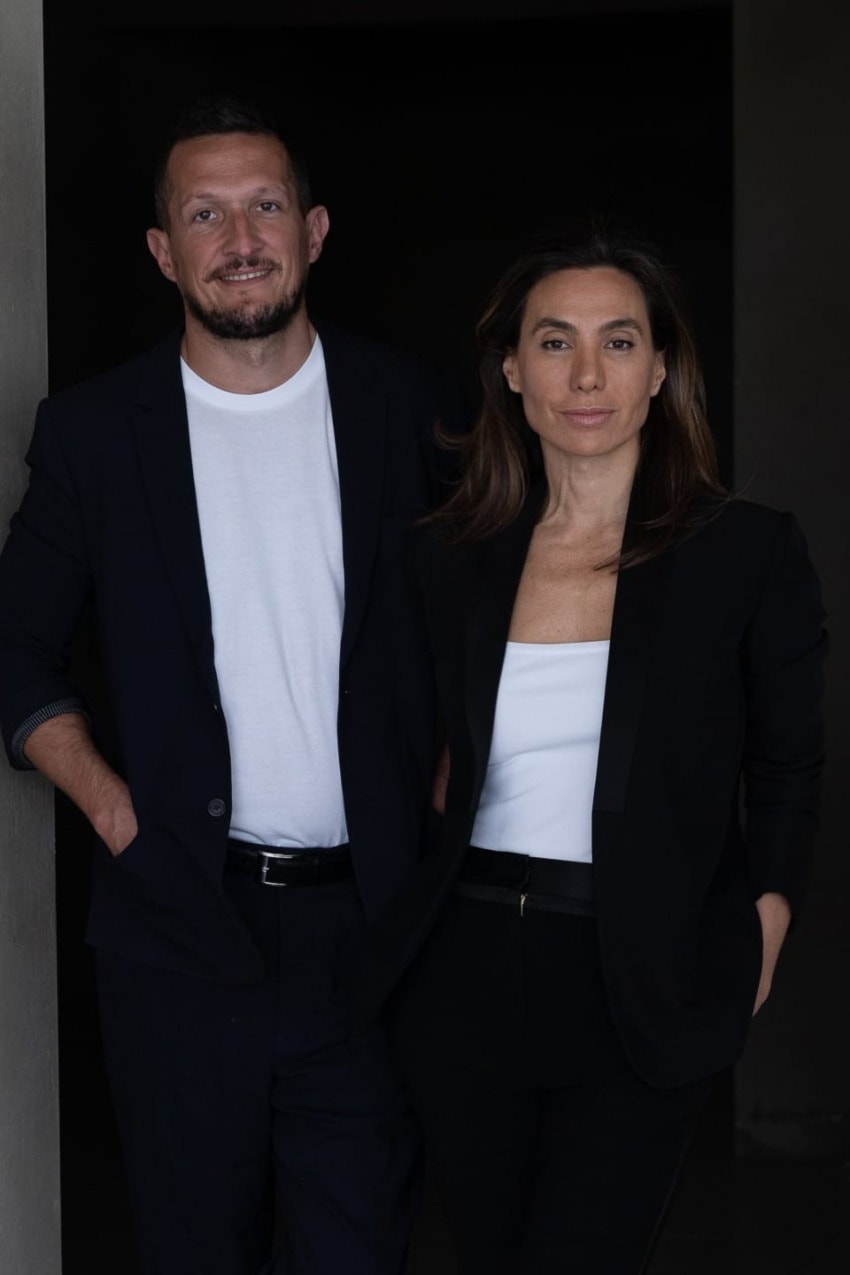 Two professionally dressed individuals standing together, the man wearing a black jacket and white shirt, the woman in a black suit.