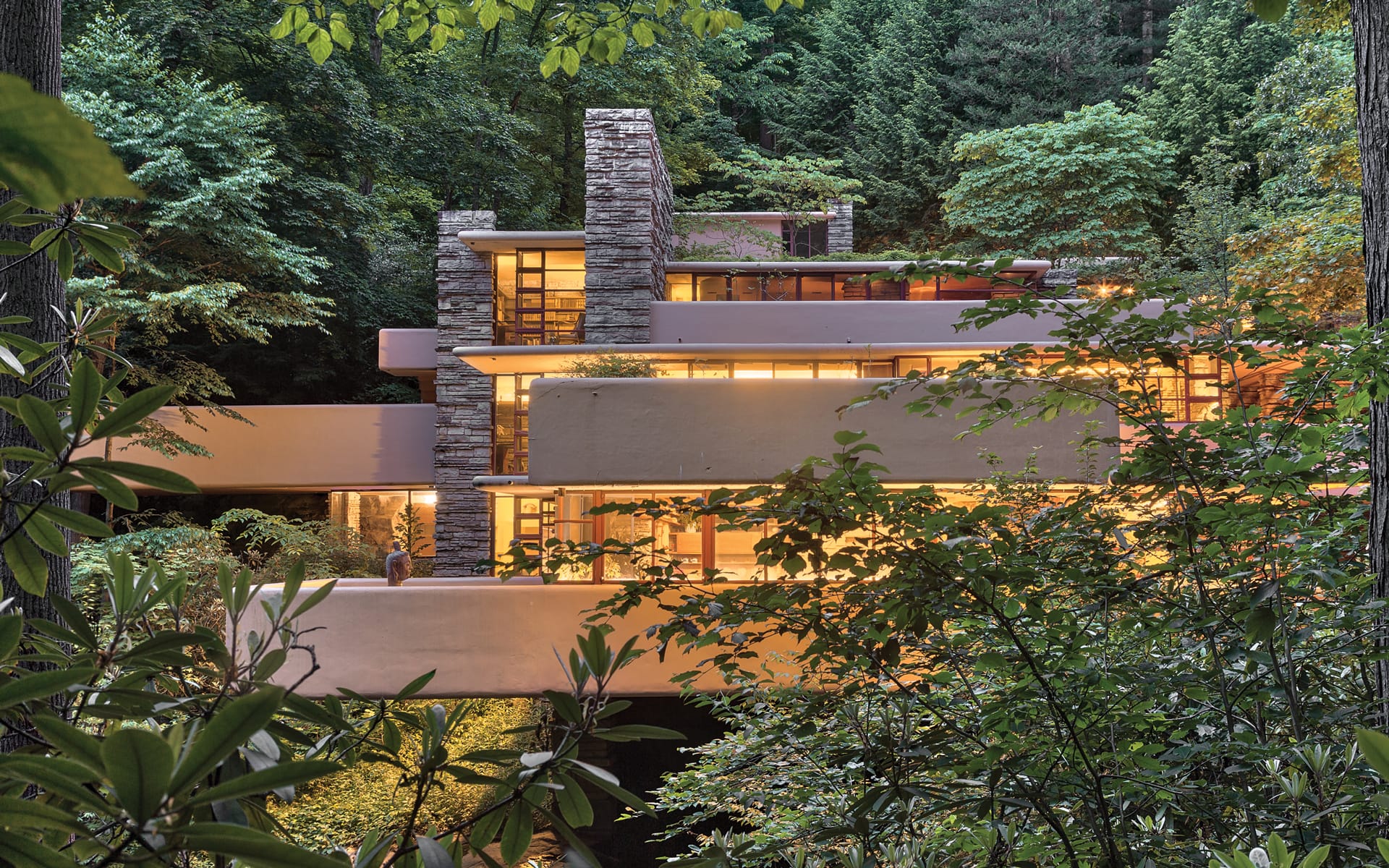Modern architectural house surrounded by lush forest and greenery, featuring stone and glass elements illuminated at dusk