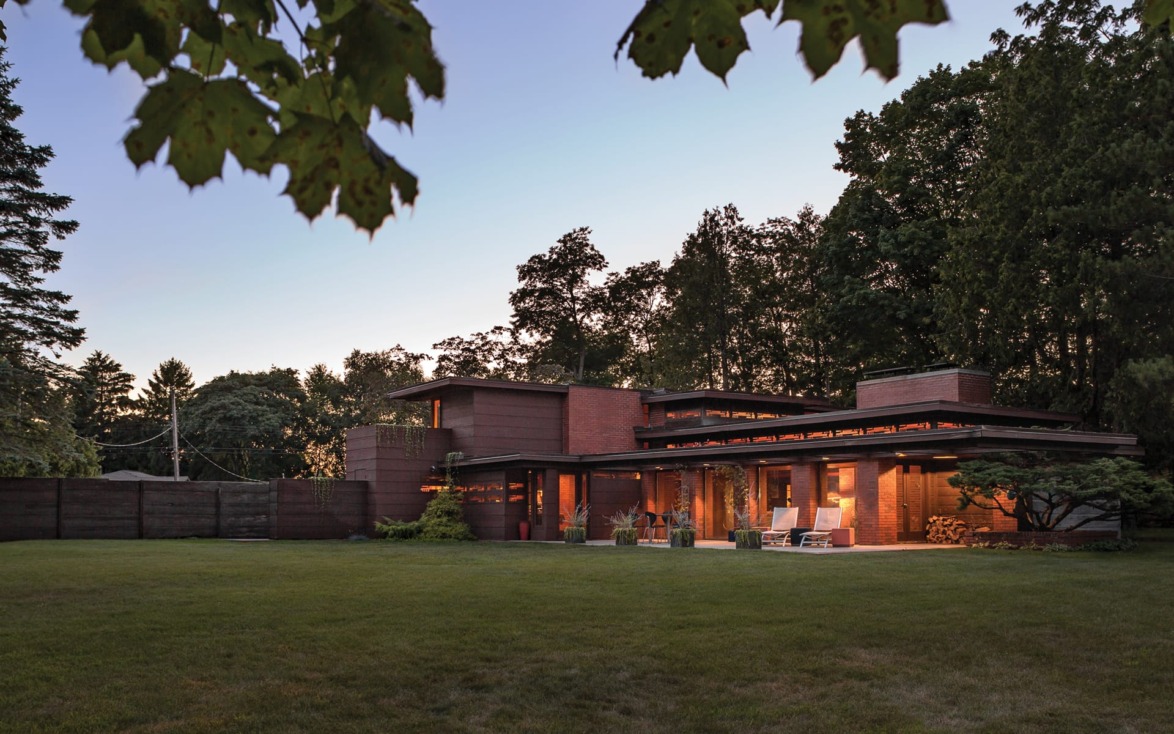 Modern brick house with large windows and a spacious lawn, surrounded by trees at dusk.