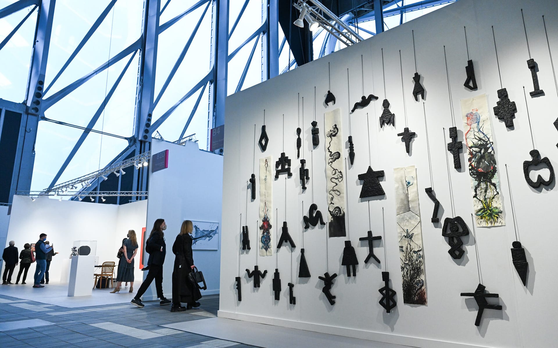 Visitors exploring a contemporary art gallery with abstract sculptures hanging on a white wall and large windows in the background.