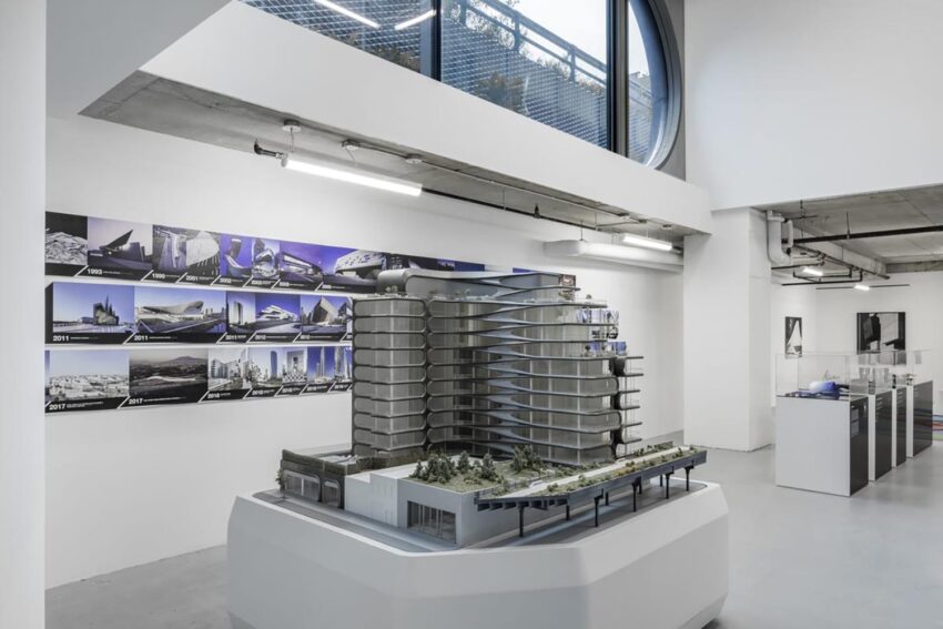 Architectural model in a modern gallery space with photographs of buildings displayed on the wall.