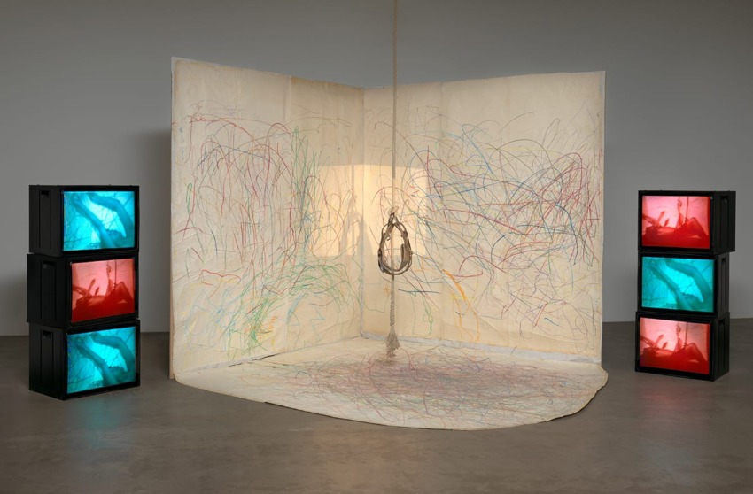 Art installation with colorful scribbles on walls and floor, surrounded by stacked TVs displaying blue and red images.