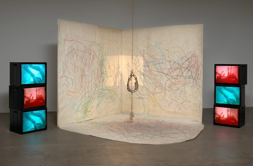 Art installation with colorful scribbles on walls and floor, surrounded by stacked TVs displaying blue and red images.