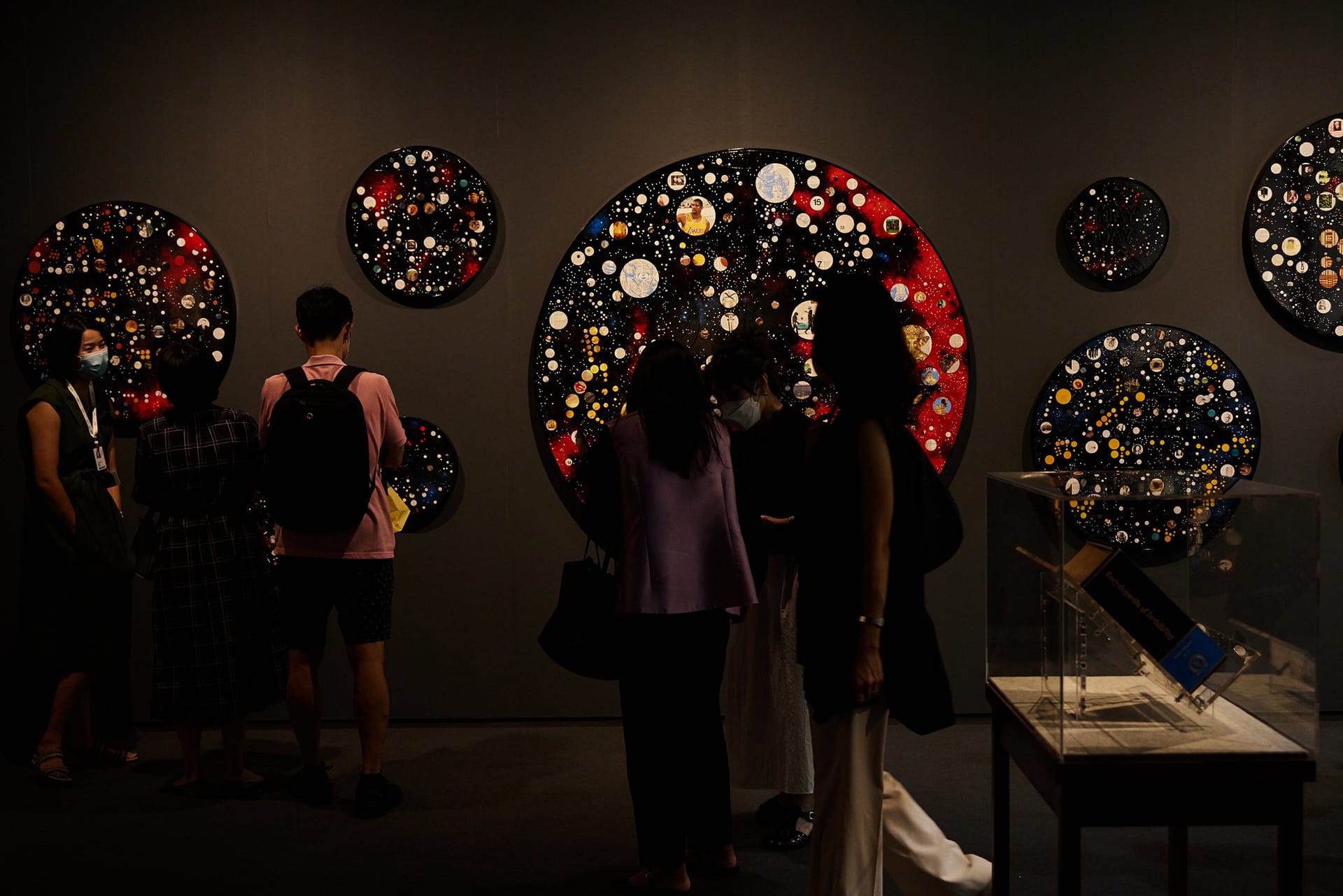 People viewing circular art pieces in dimly lit exhibition room.