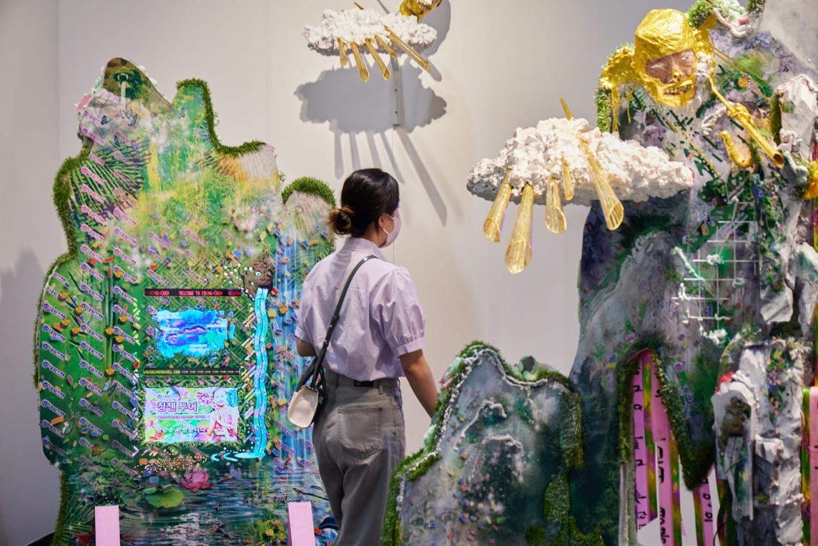 Person observing colorful, surreal art installation with clouds, greenery, and digital screen in an exhibition space.
