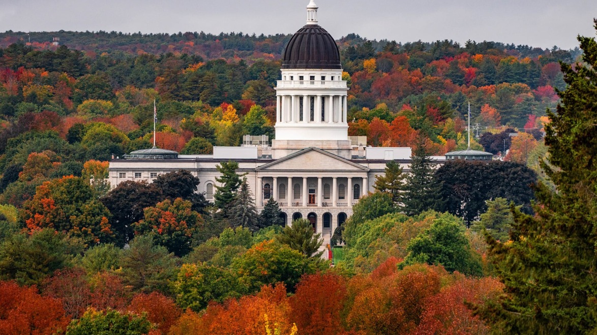 Maine State House surrounded by vibrant autumn foliage under a cloudy sky.