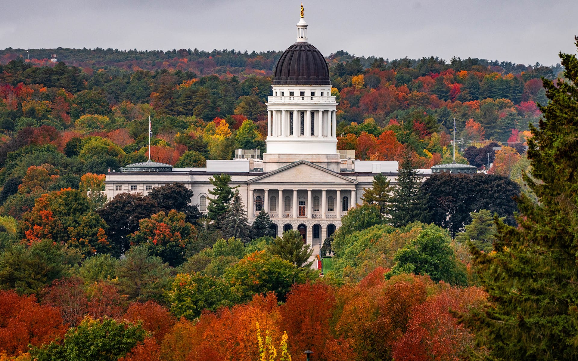 Maine State House surrounded by vibrant autumn foliage under a cloudy sky.