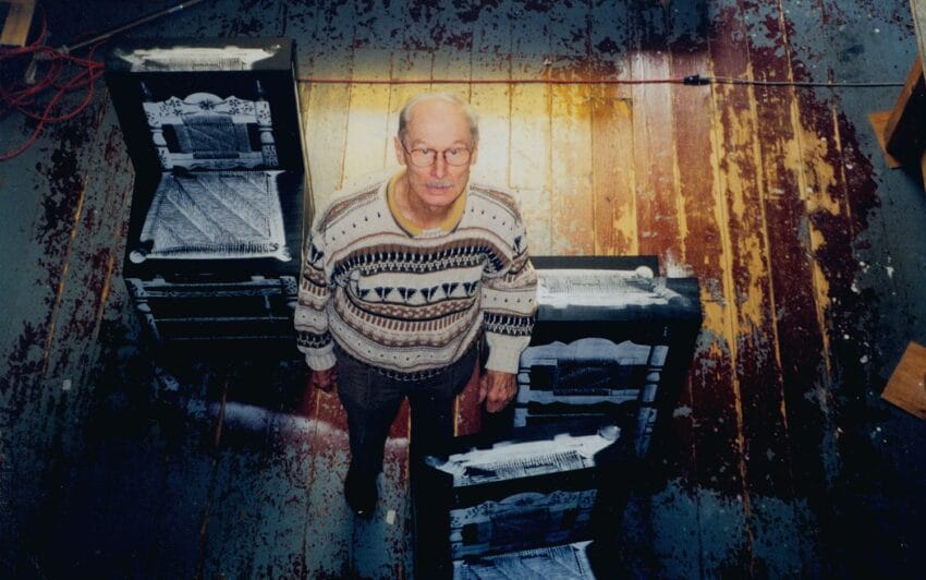 Older man in a patterned sweater stands on a worn wooden floor, surrounded by painted high-back chairs.