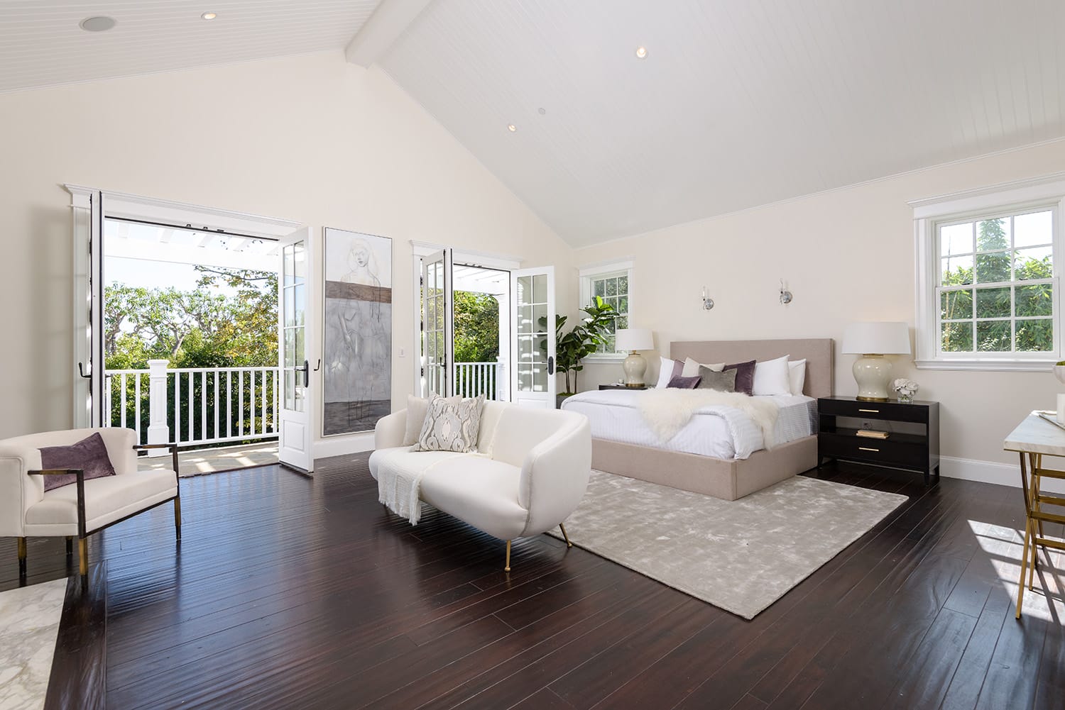 Spacious bedroom with vaulted ceiling, large bed, sofa, and open French doors leading to a balcony with a garden view.