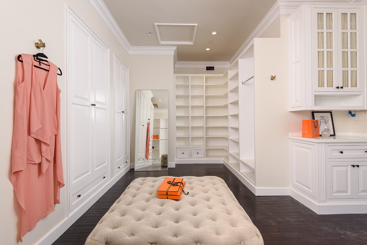 Spacious walk-in closet with white cabinets, a large mirror, pink dress hanging, and cushioned ottoman with an orange box.