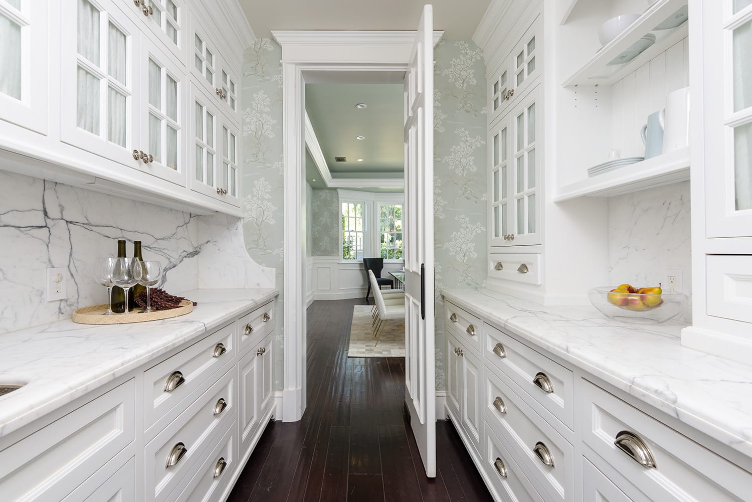 Elegant white kitchen with marble countertops, glass cabinet doors, and dark hardwood floors leading to a bright dining area.