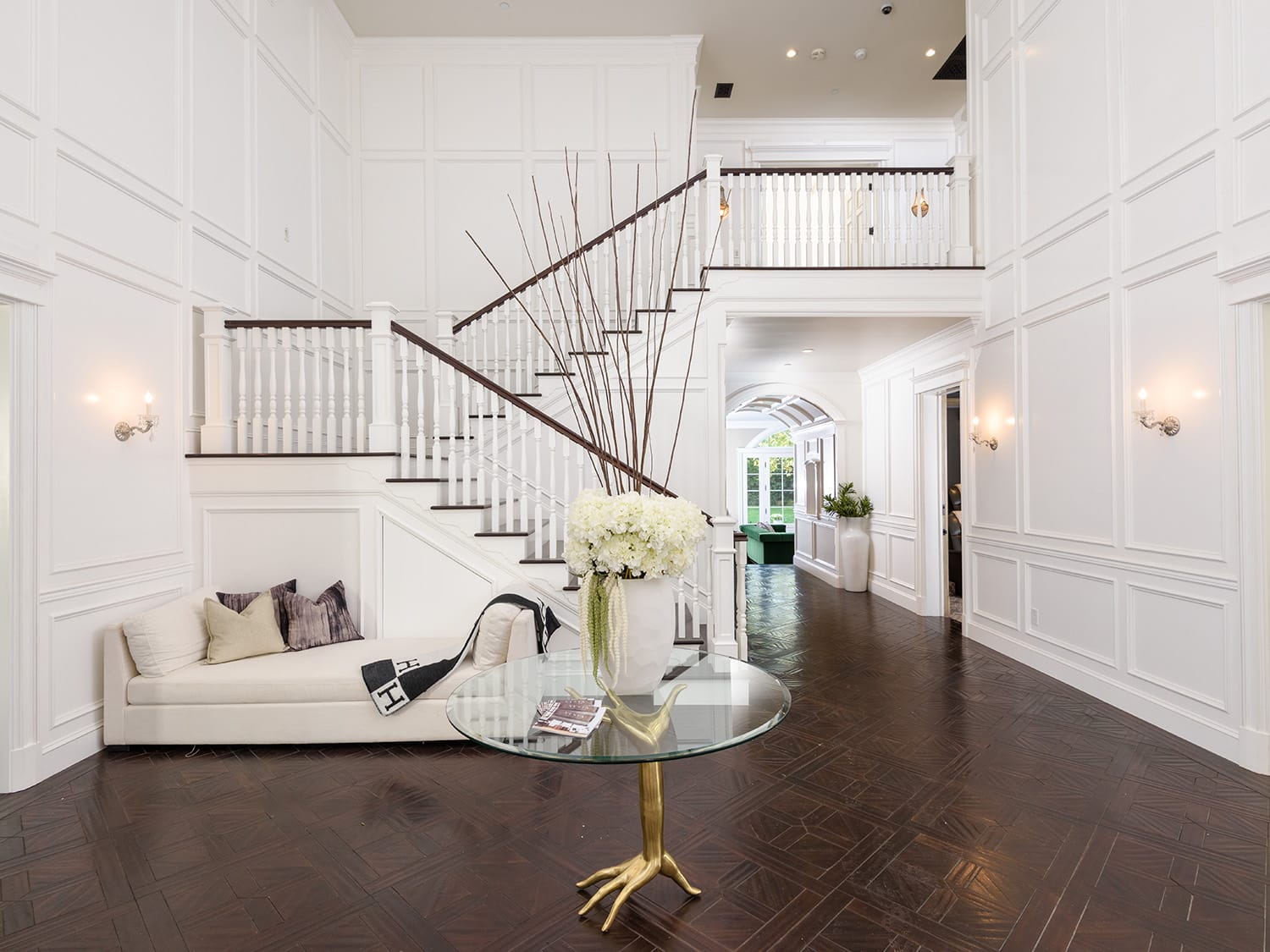 Elegant foyer with wooden staircase, white paneled walls, and dark hardwood floors featuring a round glass table and decorative vase.
