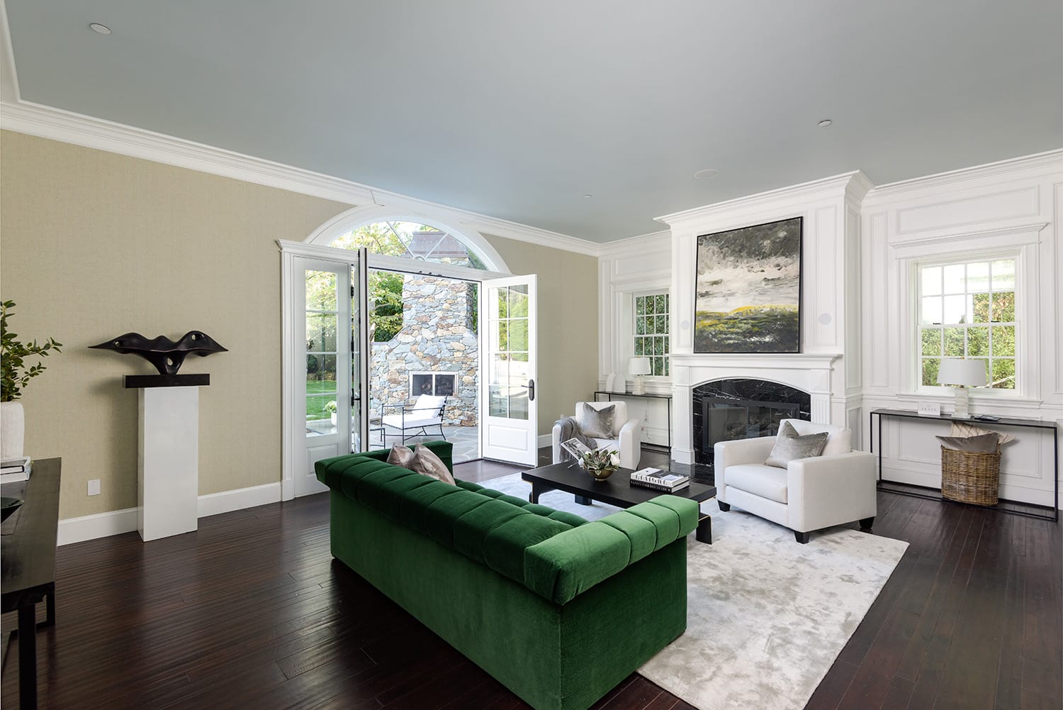Elegant living room with green sofa, white armchairs, large painting above fireplace, and view of garden through open doors.