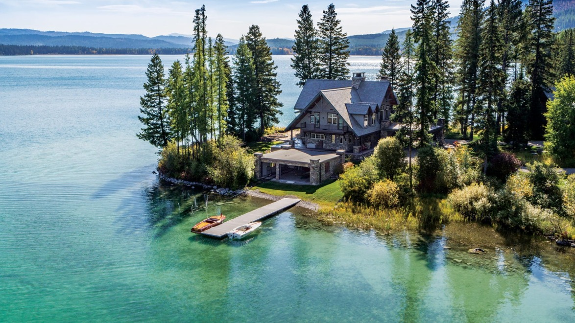 Lakeside cabin surrounded by tall trees with a dock and boats on clear, turquoise water under a blue sky.