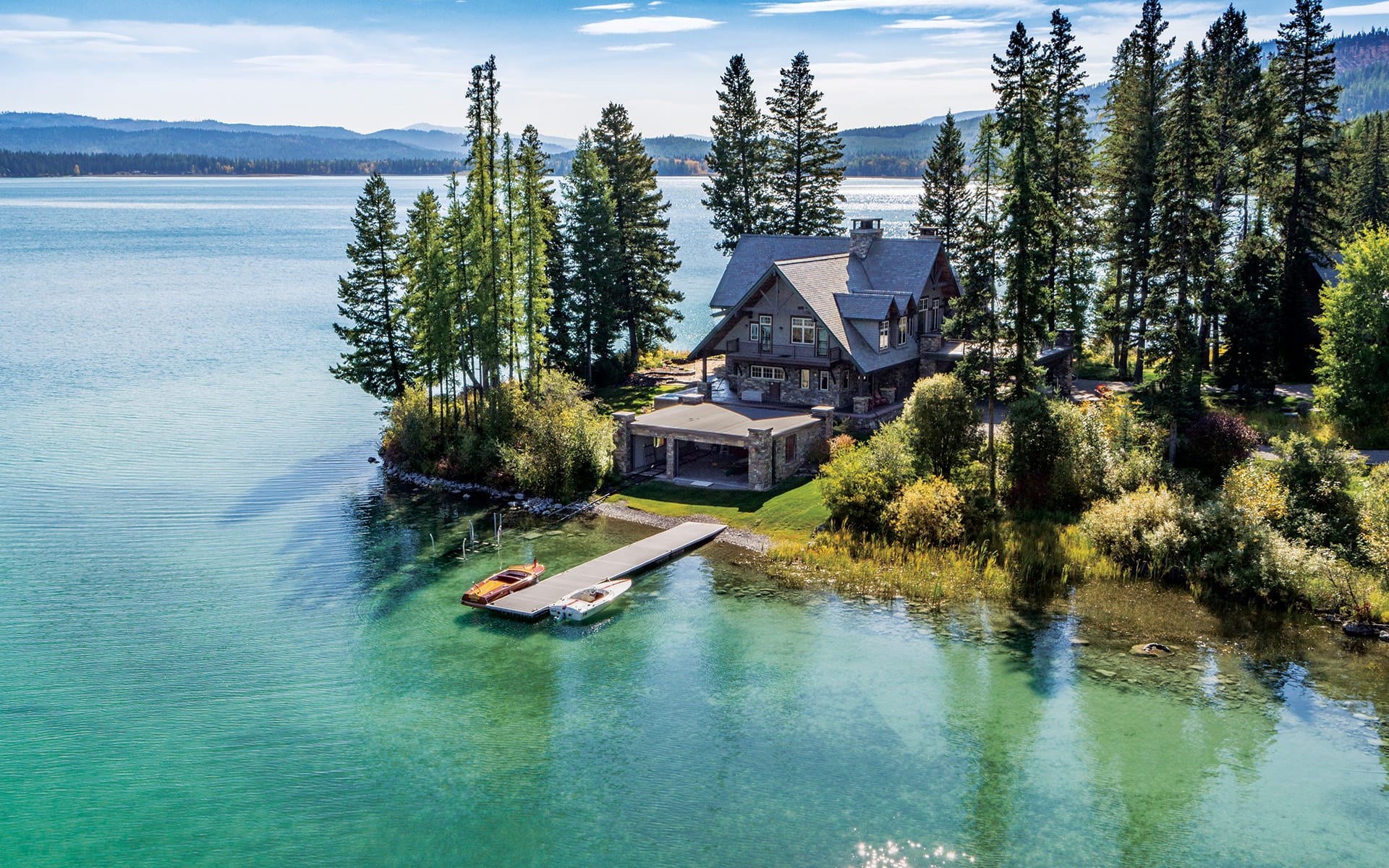 Lakeside cabin surrounded by tall trees with a dock and boats on clear, turquoise water under a blue sky.
