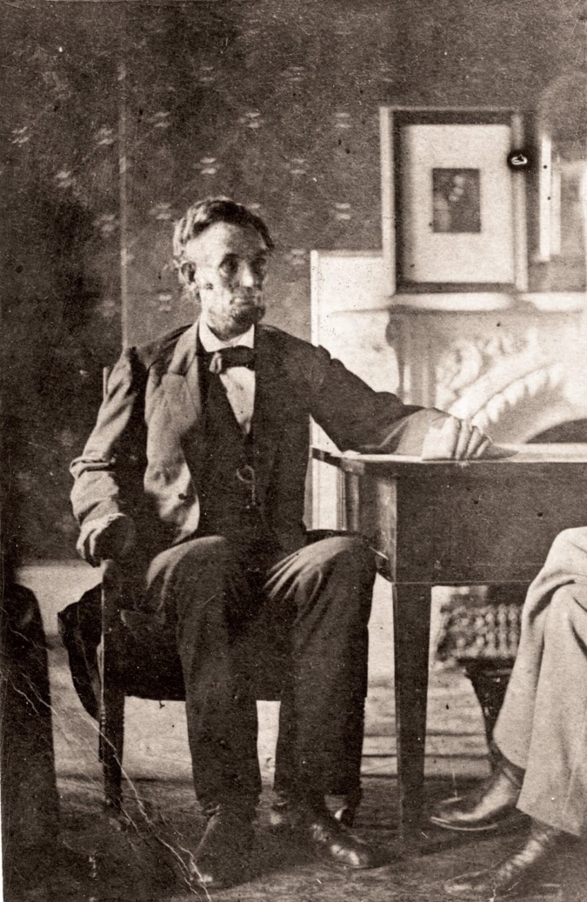 Man seated at a table wearing a suit and bow tie in a vintage sepia-toned photograph with framed pictures in the background.