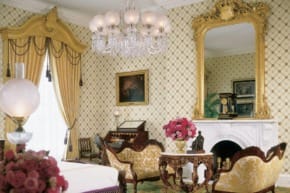 Elegant vintage living room with ornate furniture, chandelier, large mirror, floral arrangements, and patterned wallpaper.