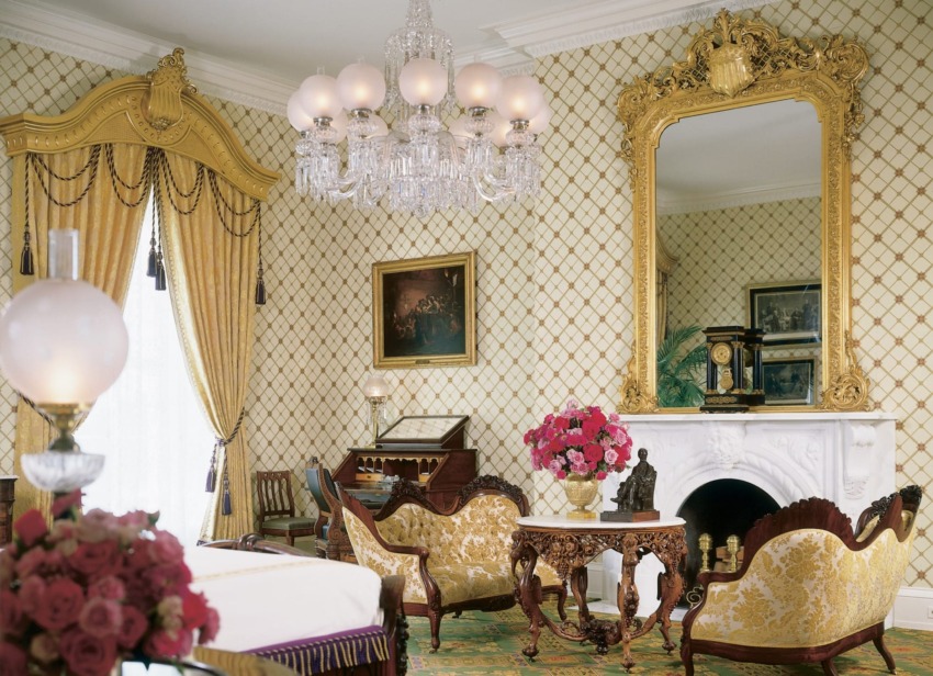 Elegant vintage living room with ornate furniture, chandelier, large mirror, floral arrangements, and patterned wallpaper.