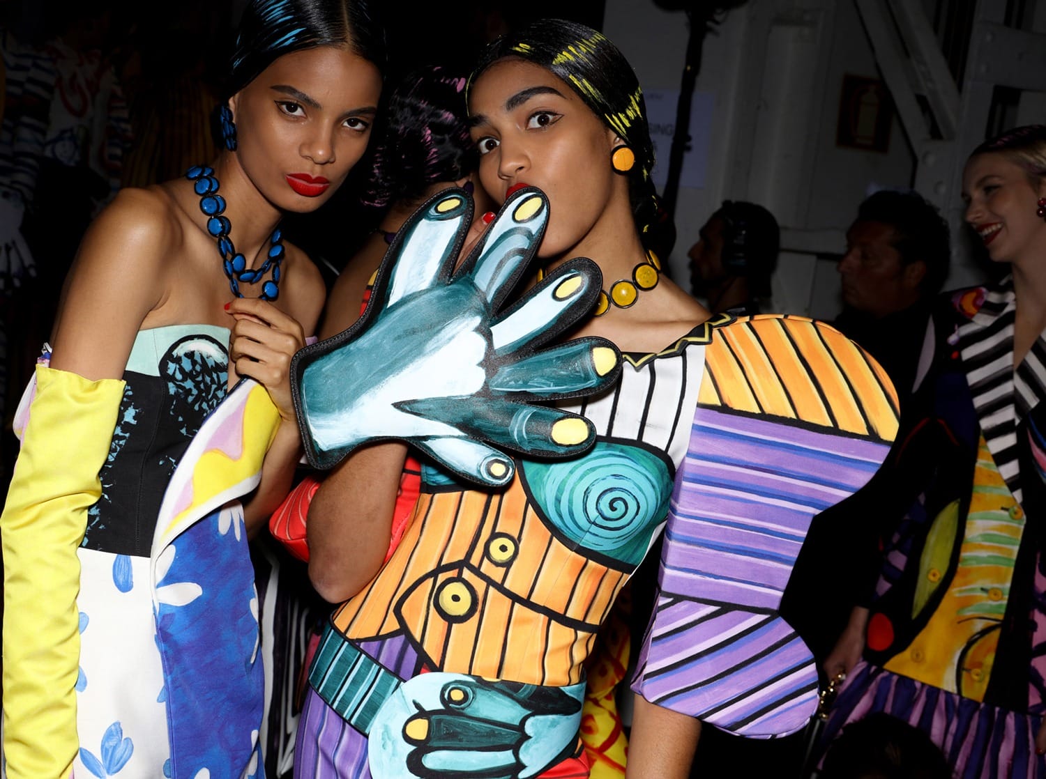 Models in vibrant, artistic outfits with bold patterns and colors pose backstage at a fashion event.